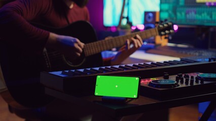 Wall Mural - Songwriter playing acoustic guitar in home studio with greenscreen display running on smartphone app. Musician practices singing on musical instrument with strings, isolated screen. Camera A.