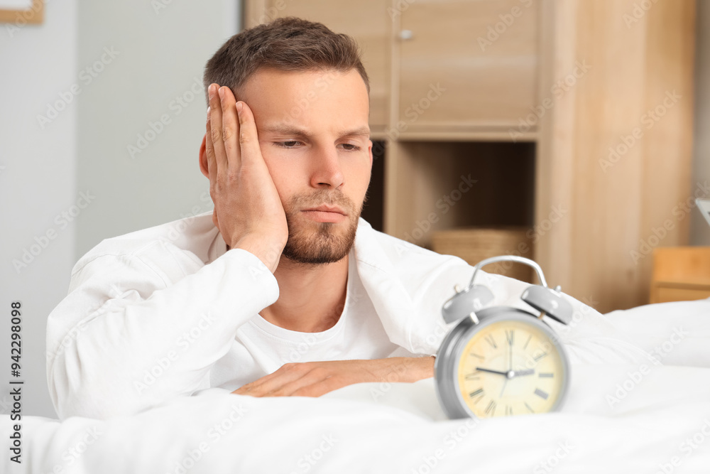 Young man and alarm clock on bed in bedroom. Insomnia concept