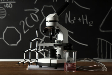 Wall Mural - Modern microscope, molecular model and flask on wooden table near chalkboard with chemical formulas