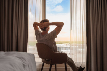 Young thoughtful man sitting back relaxing in his room hotel looking out the window 