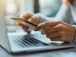 © Lightgeo - Close-up of a person's hands using a smartphone while working on a laptop, symbolizing online shopping and digital convenience.