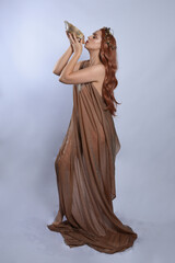 Poster - Full length portrait of beautiful female model wearing silken toga robes with golden crown, historical fantasy greek goddess aphrodite costume, holding seashell skull. Isolated on studio background