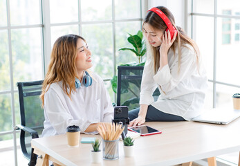 Smiling happy Asian beautiful woman sitting at table with her friend wearing wireless headphones listening music and talking, laughing in living room. Concept teenage time together lifestyle at home