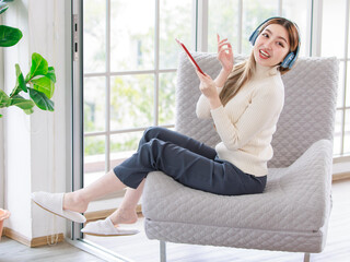 Asian young woman beautiful female happy relax listen enjoying music in headphones sitting modern sofa in front of window in living room using computer tablet. Concept of Teenage Lifestyle Hobby