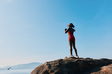 Wall Mural - Woman in red athletic wear standing on a rock by the water, with a clear blue sky in the background