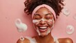© ROC WILL - A happy woman with cream/soap, isolated on a pastel pink background. A beauty portrait of a smiling African American girl, smiling while washing her face during her morning skincare routine.