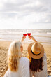 © maxbelchenko - Two woman friends enjoying red wine on a sandy beach during sunset, capturing a joyful moment filled with laughter and warmth.Hen-party.
