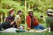 © Seventyfour - Group of friends sitting on picnic blanket in lush green park, sharing drinks and snacks, one person capturing memories with smartphone while others engage in cheerful conversation