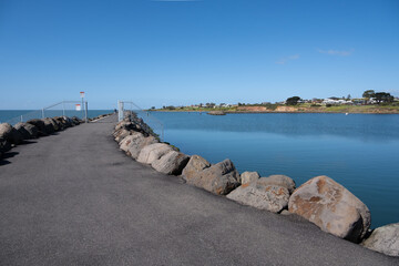 A coastal scene at Portarlington, Victoria, Australia. A paved pathway extends along a breakwater, lined with large rocks and stunning views of the calm blue ocean