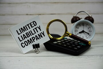 Limited Liability Company text on paper card with alarm clock, calculator and magnifying glass on wooden background