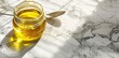© Makki - A diagonal view of a glass jar with golden oil and a spoon on a white marble table, shown closeup.
