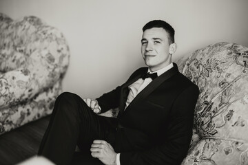 A man in a black suit and bow tie is sitting on a couch. He is smiling and he is relaxed