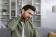 © Liubomir - Man wearing green shirt holds neck with expression of pain, sitting on sofa. Experiencing discomfort in home setting, showcasing feelings of stress and tension.