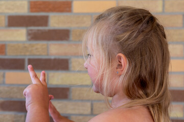 Cute chubby slightly tanned six-year old little girl with dishevelled fine blond hair seen in profile signing two with her left hand