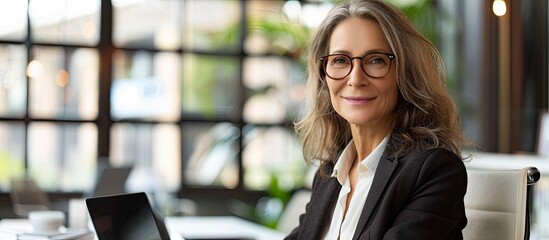 Sticker - Portrait of a happy mature businesswoman using a laptop at a desk in an office featuring bright lighting and space for additional images