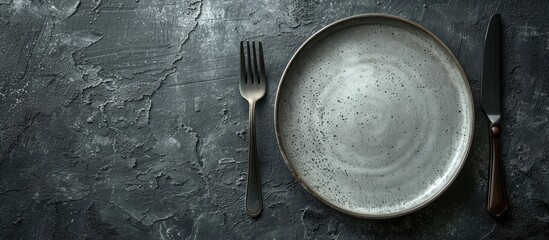 Poster - Dark textured concrete background with a round ceramic plate and cutlery setting the scene for dinner preparation with copy space image