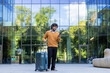 © Liubomir - Man standing with suitcase, using phone outside glass office building. Casual attire, curly hair, and smiling expression suggest successful travel or business trip.