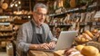 © Anastasiya - Male shop owner using a laptop in a cozy, rustic grocery store filled with bread and other products.