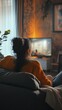 © Andrii - Cozy evening at home: person in orange sweater relaxing on comfortable couch, watching TV in warm, dimly lit living room with plants and ambient lighting.