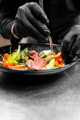 Sticker - close-up of a chef’s hands in black gloves plating a gourmet salad with sliced meat, fresh vegetables, and greens on a white plate. blurred background emphasizes the dish’s vibrant colors and textures