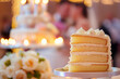 © Rohit k  - A beautifully presented slice of wedding cake sits on a plate, with a stunning tiered cake and softly lit ambiance of a wedding reception in the background. Guests enjoy the celebration