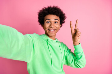 Wall Mural - Portrait of cool young man make selfie show v-sign wear green hoodie isolated on pink color background