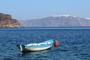 A small boat gently floats on the sparkling blue sea near Santorini, Greece. The vibrant cliffs and whitewashed buildings of the island rise in the background under the warm summer sun.