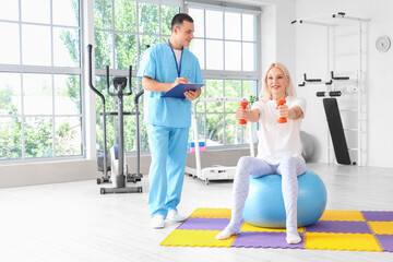 Poster - Male physiotherapist and mature woman training with dumbbells in rehabilitation center