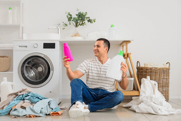 Poster - Happy young man with bottles of detergent and piles of dirty clothes sitting on floor in laundry room