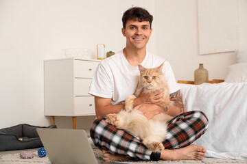 Poster - Handsome young happy man with cute Maine Coon cat and laptop sitting in bedroom at home