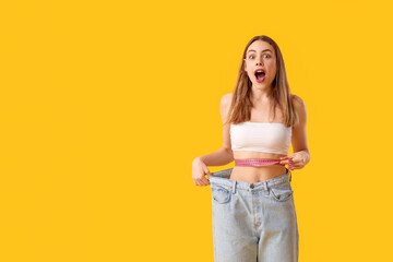 Sticker - Surprised young woman in loose jeans on yellow background. Weight loss concept