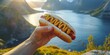 © TheWaterMeloonProjec - A hand grasping a hot dog with mustard set against a stunning natural landscape, enjoying outdoor cooking and recreational activities in the warm sunlight.