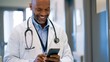 © Radomir Jovanovic - A doctor in a white coat with a stethoscope around his neck, smiling and using his smartphone in a hospital setting.