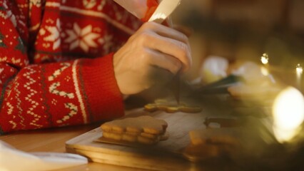 Woman in a Red Sweater Focuses on Decorating a Cookie With Red Icing, Surrounded by Other Cookies and Festive Lights at a Wooden Table.