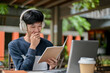 © bongkarn - A man is listening to music on his headphone while enjoying reading a book at an outdoor table.