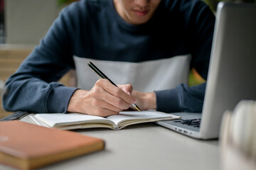 A male college student writing in his notebook or taking notes while studying online on his laptop.