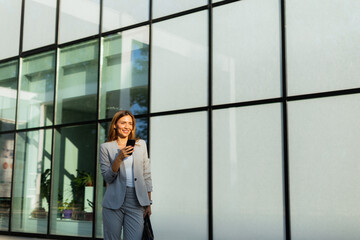 Wall Mural - Business woman strolls confidently outdoors in smart attire, checking her messages against the backdrop of a modern glass building on a sunny day