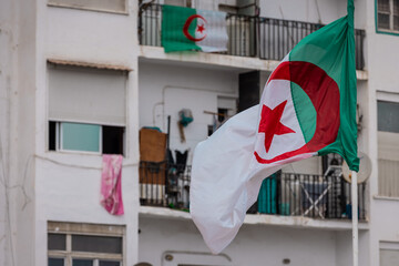 Flag of Algeria waving in a city environment, behind apartment building with balconies and satellite dishes. Poverty in Algeria.