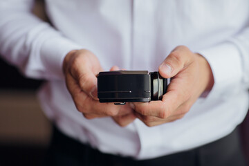 A man is holding a camera in his hand. The camera is black and has a square shape. The man is wearing a white shirt and black pants. Concept of professionalism and focus