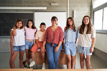 Cheerful portrait of a group of preteen students with their female teacher inside the classroom looking at camera smiling, enjoying their back to school.