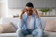 © Home-stock - Stressed young black man sitting on couch at home, rubbing temples, suffering from headache migraine or thinking