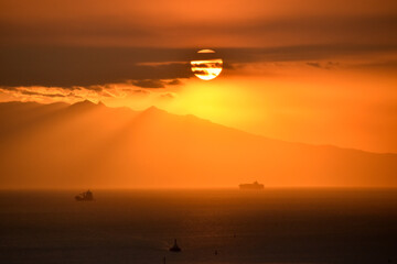 Highly dramatic and cinematic view with a silhouette of a mountain and two boats at sea casting an orange hue over a calm sea