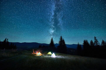 Blurred silhouettes of tourists resting at campsite around campfire with glowing tents nearby under starry sky. Milky Way stretches above, casting magical light over tranquil mountain landscape.