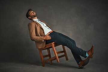 Handsome man, laughing and leaning with chair for funny humor, joke or comedy on a gray background. Young, male person or model with smile in laughter for stylish fashion, retro or vintage outfit