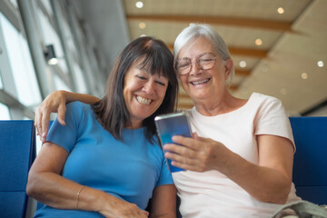 Sticker - Smiling senior couple of female friends sitting in airport departure area waiting for boarding looking at mobile phone. Travel and tourism concept, retiree lifestyle