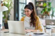 © miss irine - Young woman sits at desk with laptop. Concentrated and smiling, she works in comfortable casual attire. Beautiful background with bright light, highlighting her confident expression.