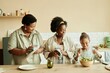 © Seventyfour - Medium shot of African American mother and granny wiping dishes at large wooden counter watching smiling girl making fresh salad at cozy beige kitchen