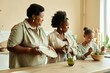 © Seventyfour - Side view of smiling African American mother and granny wiping dishes at large wooden counter watching little girl making fresh salad at cozy beige kitchen