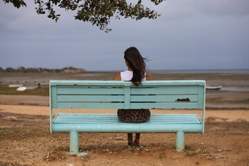 Wall Mural - beautiful young Spanish woman sitting on her back on a light blue bench looking at the horizon. The day is cloudy and grey and you can see boats stranded on the shore of the bay of Cadiz, Spain.