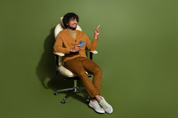 Sticker - Full length photo of handsome young guy sit armchair device point look empty space dressed brown garment isolated on khaki color background